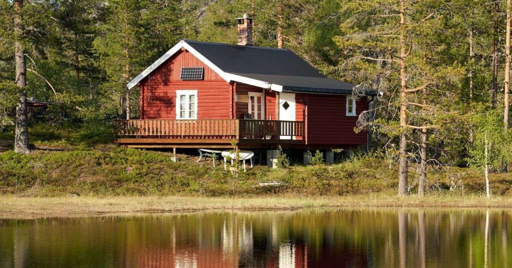 Punainen mökki tyynen järven rannalla. Mökin ympärillä metsää.